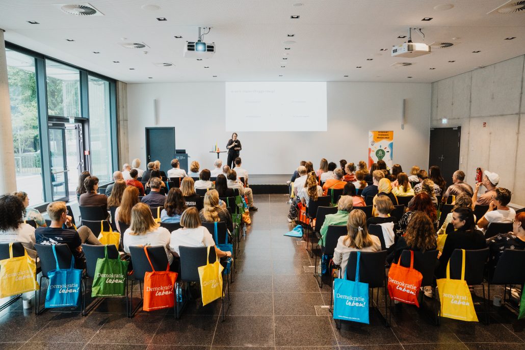 Ein größerer Seminarraum mit vielen Teilnehmenden, die in Reihen sitzen und einer Vortragenden auf einer Bühne zuhören. Auf den Stühlen hängen bunte Stofftaschen mit der Aufschrift ‚Demokratie Leben‘. Auf der Bühne steht eine Person vor einer Präsentationsfolie, die an die Wand projiziert wird. Rechts daneben befindet sich ein farbiges Plakat. Durch die großen Fenster links fällt Tageslicht in den Raum.