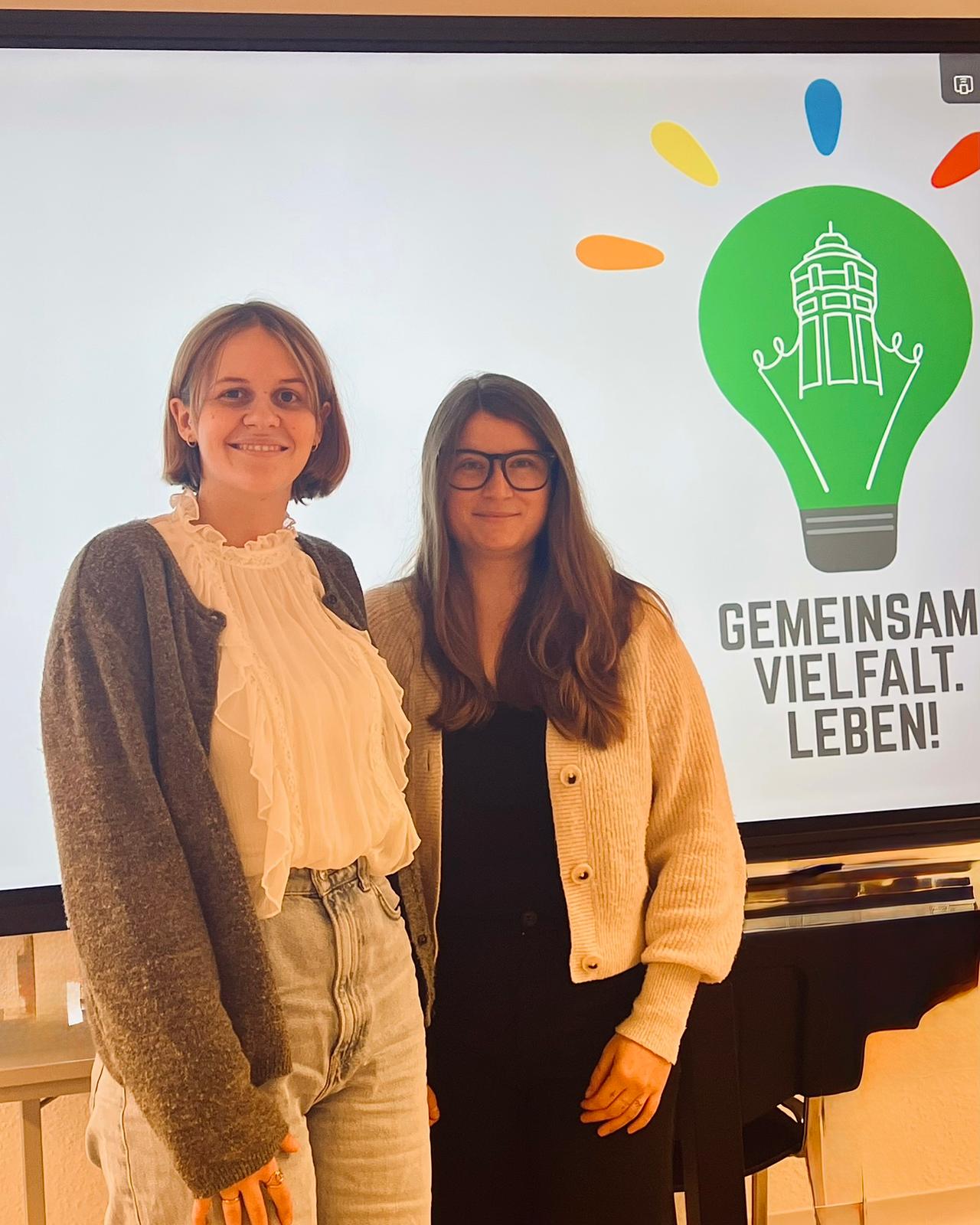 Zwei Personen stehen in einem Raum vor einem großen Bildschirm. Auf dem Bildschirm ist eine grüne Glühbirne mit stilisiertem Gebäude sowie farbige Akzente zu sehen, daneben der Schriftzug „Gemeinsam Vielfalt. Leben!“. Die beiden Personen tragen helle Strickjacken und stehen nebeneinander im Vordergrund.