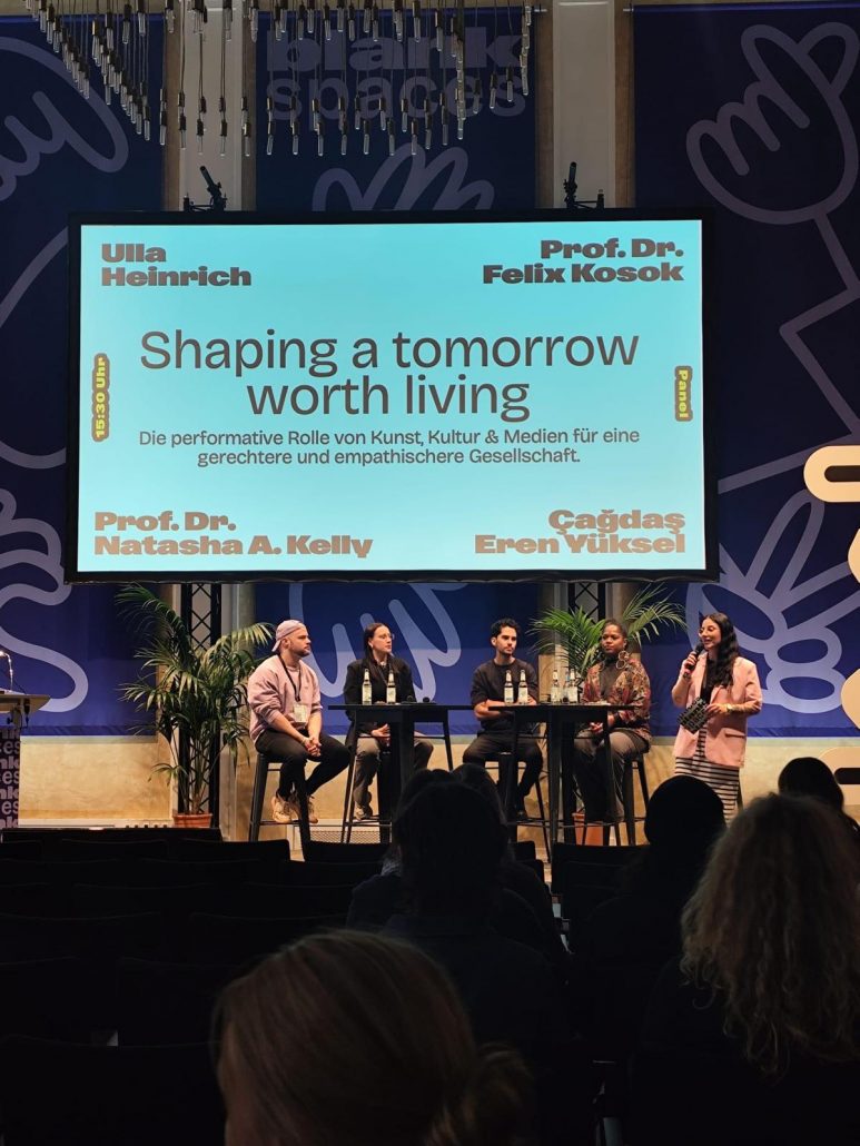Auf einer Bühne sitzen fünf Personen an einem hohen Tisch in einer Paneldiskussion. Hinter ihnen hängt eine große Leinwand mit dem Titel „Shaping a tomorrow worth living“ und dem Untertitel „Die performative Rolle von Kunst, Kultur & Medien für eine gerechtere und empathischere Gesellschaft“. Auf der Leinwand stehen außerdem die Namen der beteiligten Sprecherinnen und Sprecher. Die Bühne ist von Pflanzen eingerahmt, und im Publikum sitzen mehrere Personen, die die Veranstaltung verfolgen.