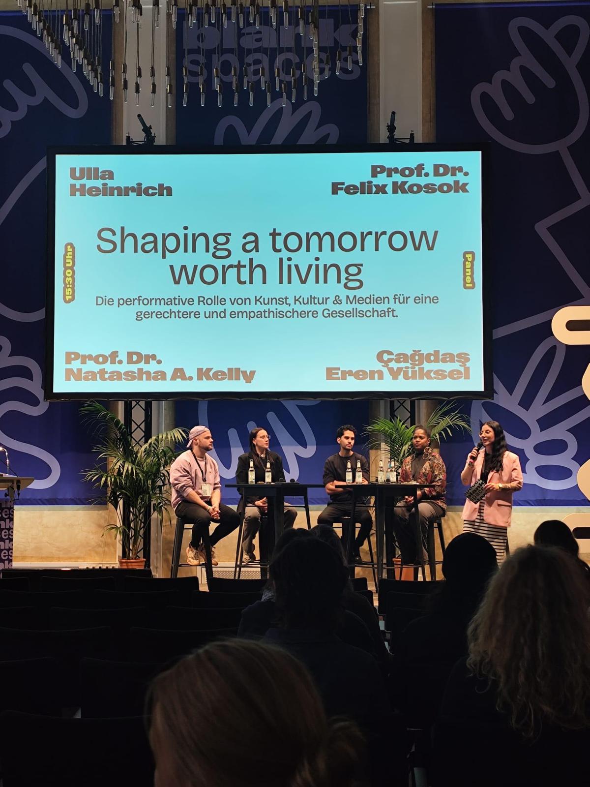 Auf einer Bühne sitzen fünf Personen an einem hohen Tisch in einer Paneldiskussion. Hinter ihnen hängt eine große Leinwand mit dem Titel „Shaping a tomorrow worth living“ und dem Untertitel „Die performative Rolle von Kunst, Kultur & Medien für eine gerechtere und empathischere Gesellschaft“. Auf der Leinwand stehen außerdem die Namen der beteiligten Sprecherinnen und Sprecher. Die Bühne ist von Pflanzen eingerahmt, und im Publikum sitzen mehrere Personen, die die Veranstaltung verfolgen.