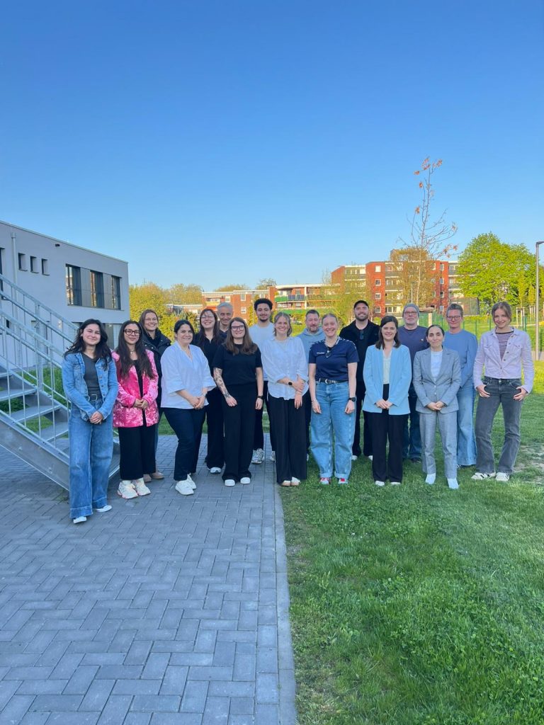 Gruppenfoto von Teilnehmenden im Außenbereich eines Bildungsstandorts vor modernen Gebäuden an einem sonnigen Tag.