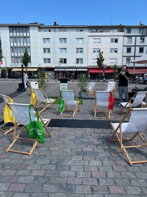 Auf dem Rheydter Markt ist die Veranstaltung ‚Wie du hineinrufst‘ aufgebaut. Zu sehen sind mehrere Liegestühle, bepflanzte Elemente, Lautsprecher und ein Redepult. An den Liegestühlen hängen grüne und gelbe Stoffbeutel mit dem Logo von ‚Demokratie leben!‘. Im Hintergrund befinden sich Gebäude und weitere Bereiche des Marktplatzes