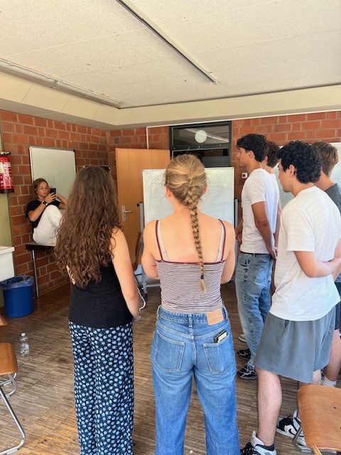 Mehrere Jugendliche arbeiten während eines Workshops gemeinsam an einer Aufgabenstellung in einem Seminarraum. Die Teilnehmenden stehen vor einem Whiteboard und tauschen sich in Kleingruppen aus. Der Raum ist mit Stühlen, Tischen und Moderationsmaterial ausgestattet.
